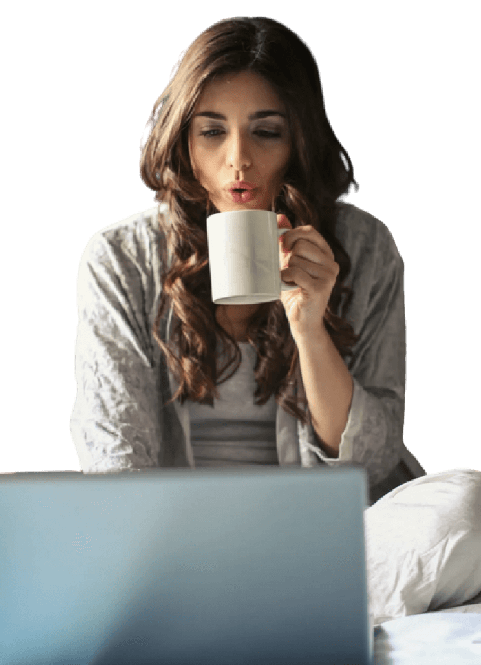 Woman with long brown hair blowing on a white mug while sitting at a laptop.